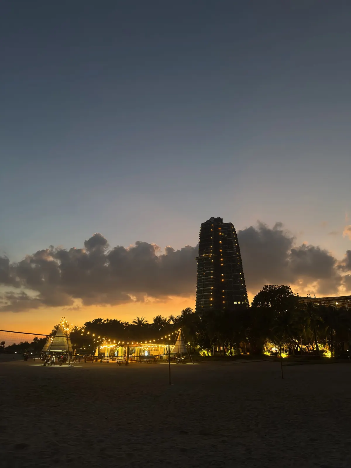 Sunset over a sandy beach, a tall tower silhouetted against an orange horizon, strings of lights among the palms.