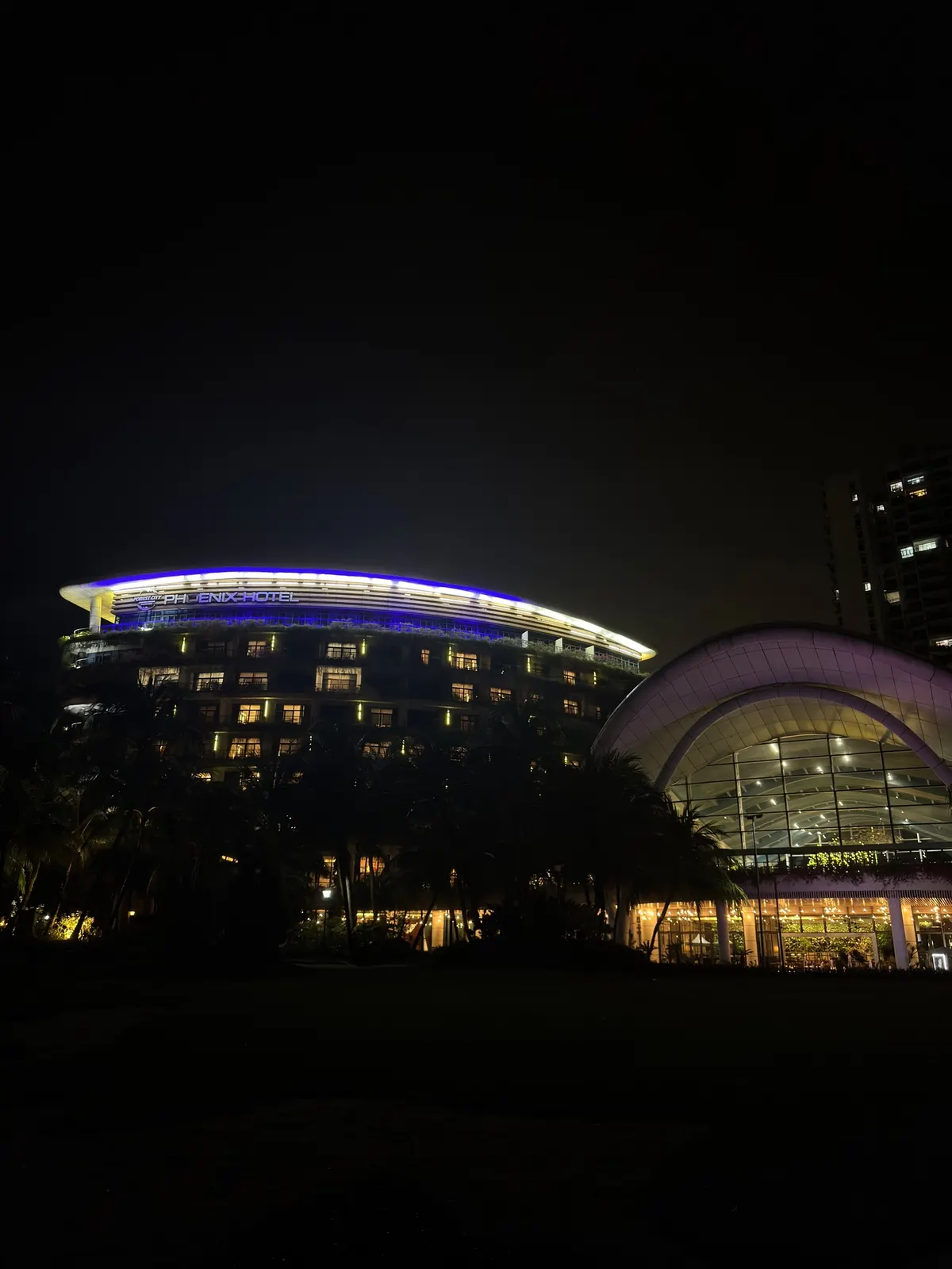 A curved waterfront building lit at night beside a glass dome glowing pink and gold, dark lawns in the foreground.