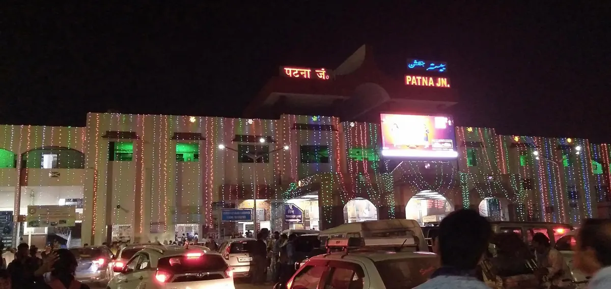 Patna Junction railway station at night, its facade strung with coloured lights and a lit sign, cars and people in the forecourt.