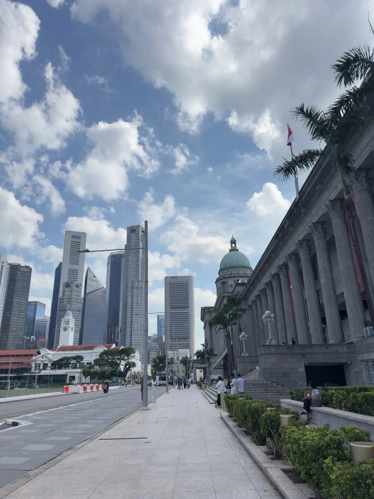 A wide pavement beside a colonnaded colonial facade, the green dome of the National Gallery and the towers of the business district beyond, under a blue sky with cumulus clouds.